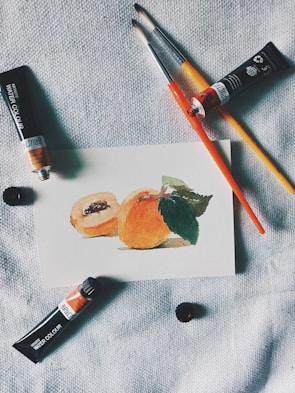 A small watercolor painting of peaches rests on a textured fabric surface. Nearby are paint brushes and tubes of watercolor paint, arranged casually around the artwork, suggesting an artist's workspace.