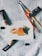 A small watercolor painting of peaches rests on a textured fabric surface. Nearby are paint brushes and tubes of watercolor paint, arranged casually around the artwork, suggesting an artist's workspace.