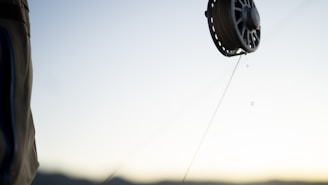 A close-up of a fishing reel with a line extending from it, set against a blurred background of a clear sky and distant mountains. The focus on the reel captures fine details as droplets of water hang from the line.