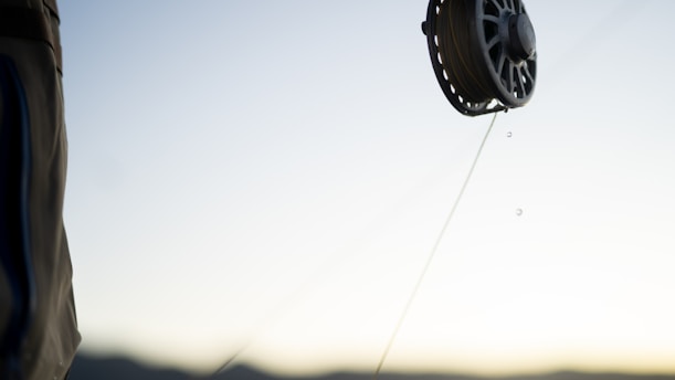 A close-up of a fishing reel with a line extending from it, set against a blurred background of a clear sky and distant mountains. The focus on the reel captures fine details as droplets of water hang from the line.
