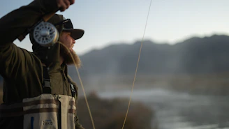 Angler casting a fly rod over a serene mountain stream surrounded by pine trees.