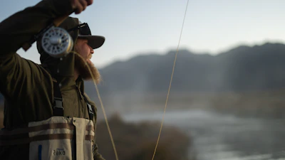 Angler casting a fly rod over a serene mountain stream surrounded by pine trees.