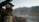 A person dressed in outdoor gear with a beard is fly fishing beside a river. They hold a fishing rod and reel, casting the line with mountains in the background. The scene is serene and focused on the angler's activity.