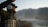 A person dressed in outdoor gear with a beard is fly fishing beside a river. They hold a fishing rod and reel, casting the line with mountains in the background. The scene is serene and focused on the angler's activity.
