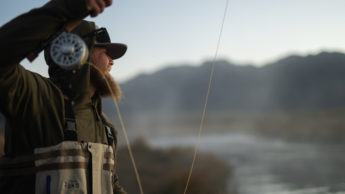 A person dressed in outdoor gear with a beard is fly fishing beside a river. They hold a fishing rod and reel, casting the line with mountains in the background. The scene is serene and focused on the angler's activity.