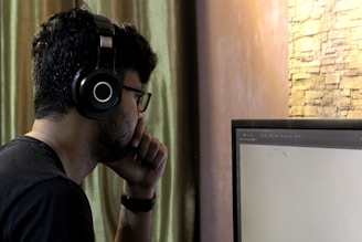 A person wearing black headphones and glasses sits in front of a computer monitor. The individual is resting their chin on their hand, appearing focused and engaged with the screen. The background features a curtain and a stone-textured wall.