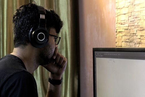 A person wearing black headphones and glasses sits in front of a computer monitor. The individual is resting their chin on their hand, appearing focused and engaged with the screen. The background features a curtain and a stone-textured wall.