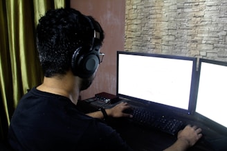 A person wearing headphones sits in front of a computer setup with two monitors illuminated in a dimly lit room. The walls have a textured stone finish, and the curtain is a dark green color. The atmosphere suggests focus and concentration.