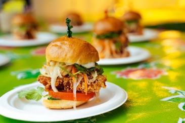 A close-up view of a gourmet sandwich featuring a crispy fried chicken patty topped with fresh vegetables, such as shredded lettuce and carrots, placed in a soft, glossy bun. The sandwich is elegantly presented on a white plate, with a vibrant, colorful background enhancing its appeal.