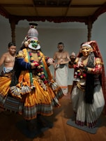 A traditional dance scene features two performers in elaborate costumes and makeup, showcasing vibrant colors and intricate designs. Two onlookers in simple attire stand in the background, adding depth to the cultural setting. The foreground costumes are detailed with golden yellow fabric, ornate jewelry, and painted faces, indicating a classical theatrical performance.