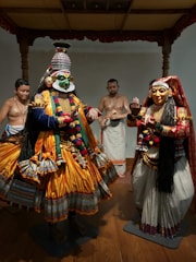 A traditional dance scene features two performers in elaborate costumes and makeup, showcasing vibrant colors and intricate designs. Two onlookers in simple attire stand in the background, adding depth to the cultural setting. The foreground costumes are detailed with golden yellow fabric, ornate jewelry, and painted faces, indicating a classical theatrical performance.