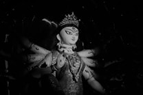 A monochrome image of a statue depicting a deity with multiple arms, each positioned differently. The figure has intricately detailed clothing with elaborate patterns, and its head is adorned with a crown. The facial expression is serene and focused, conveying a sense of peace and strength.