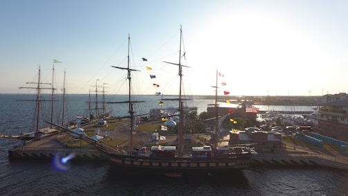 A large sailing ship docked at a port, adorned with colorful flags. The sun is setting in the background, casting a warm glow over the scene. Several buildings and additional ships are visible, with the ocean stretching out to the horizon.