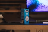 A box of Durex Classic condoms is placed on a wooden table. In the background, there is a blurred image of a computer screen displaying a blue and purple pattern. A portion of a keyboard or remote is visible on the table next to the box. The setting appears to be indoors with a dim, ambient lighting.