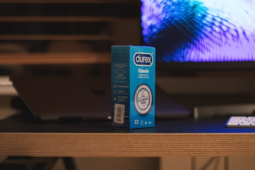 A box of Durex Classic condoms is placed on a wooden table. In the background, there is a blurred image of a computer screen displaying a blue and purple pattern. A portion of a keyboard or remote is visible on the table next to the box. The setting appears to be indoors with a dim, ambient lighting.