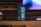 A box of Durex Classic condoms is placed on a wooden table. In the background, there is a blurred image of a computer screen displaying a blue and purple pattern. A portion of a keyboard or remote is visible on the table next to the box. The setting appears to be indoors with a dim, ambient lighting.