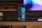 A box of Durex Classic condoms is placed on a wooden table. In the background, there is a blurred image of a computer screen displaying a blue and purple pattern. A portion of a keyboard or remote is visible on the table next to the box. The setting appears to be indoors with a dim, ambient lighting.