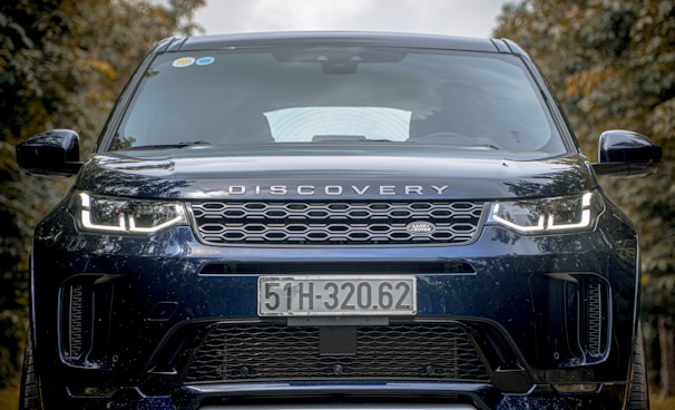 A close-up view of the front of a black Land Rover Discovery vehicle with a distinct grille and prominent headlights. The license plate displays the number 51H-320.62, and there are reflections visible on the windshield. The background consists of blurred trees, suggesting an outdoor setting.