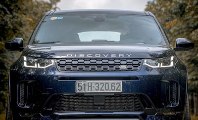 A close-up view of the front of a black Land Rover Discovery vehicle with a distinct grille and prominent headlights. The license plate displays the number 51H-320.62, and there are reflections visible on the windshield. The background consists of blurred trees, suggesting an outdoor setting.