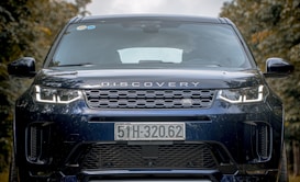 A close-up view of the front of a black Land Rover Discovery vehicle with a distinct grille and prominent headlights. The license plate displays the number 51H-320.62, and there are reflections visible on the windshield. The background consists of blurred trees, suggesting an outdoor setting.