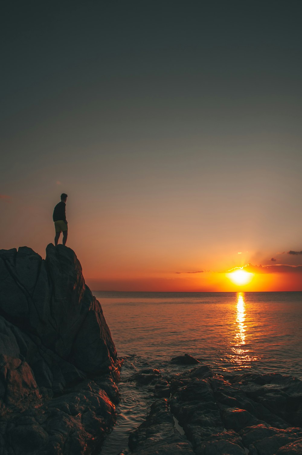 Silhouette d’une personne debout sur une formation rocheuse près d’un plan d’eau au coucher du soleil