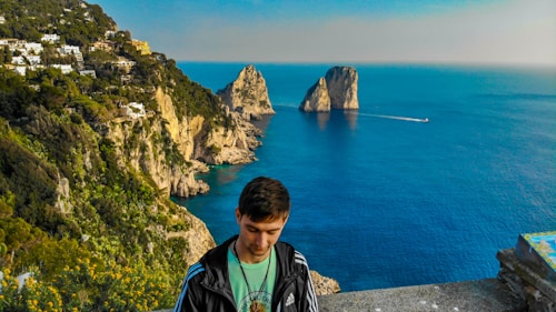 A young person stands in the foreground overlooking a breathtaking coastal landscape. The scene includes steep, rocky cliffs adorned with lush greenery and scattered buildings. The deep blue sea stretches out to the horizon, punctuated by two prominent rock formations. A boat creates a white trail across the serene water.