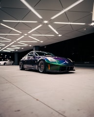 A sleek sports car with a vibrant chameleon paint finish is parked under a geometrically patterned ceiling with bright lighting. The car's dynamic design is complemented by custom black rims and low-profile tires. In the background, there is another vehicle, partially visible.