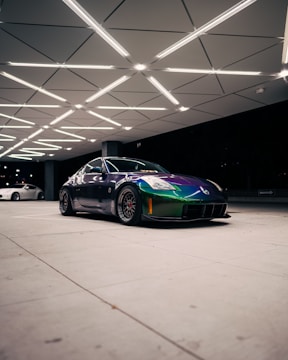 A sleek sports car with a vibrant chameleon paint finish is parked under a geometrically patterned ceiling with bright lighting. The car's dynamic design is complemented by custom black rims and low-profile tires. In the background, there is another vehicle, partially visible.