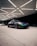 A sleek sports car with a vibrant chameleon paint finish is parked under a geometrically patterned ceiling with bright lighting. The car's dynamic design is complemented by custom black rims and low-profile tires. In the background, there is another vehicle, partially visible.