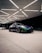 A sleek sports car with a vibrant chameleon paint finish is parked under a geometrically patterned ceiling with bright lighting. The car's dynamic design is complemented by custom black rims and low-profile tires. In the background, there is another vehicle, partially visible.