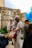 A group of people gathered in an outdoor setting with stone walls. A man is playing a drum, and another is playing a saxophone. Blue and white balloons are visible, and some people are wearing hats and traditional clothing.