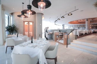 A spacious and modern restaurant interior with neatly arranged tables and chairs. The tables are set with white tablecloths, glasses, and cutlery. The lighting is soft and warm, creating a welcoming atmosphere. There are a few large pendant lights hanging from the ceiling, and the floor is made of polished marble. In the background, a couple of restaurant staff members are visible, adding to the lively yet refined environment.