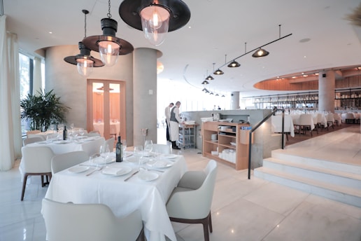 A spacious and modern restaurant interior with neatly arranged tables and chairs. The tables are set with white tablecloths, glasses, and cutlery. The lighting is soft and warm, creating a welcoming atmosphere. There are a few large pendant lights hanging from the ceiling, and the floor is made of polished marble. In the background, a couple of restaurant staff members are visible, adding to the lively yet refined environment.