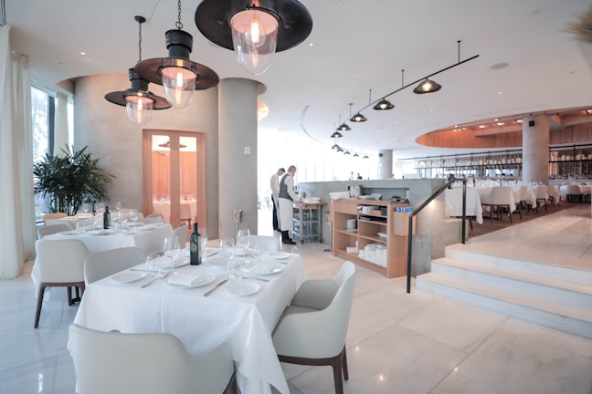 A spacious and modern restaurant interior with neatly arranged tables and chairs. The tables are set with white tablecloths, glasses, and cutlery. The lighting is soft and warm, creating a welcoming atmosphere. There are a few large pendant lights hanging from the ceiling, and the floor is made of polished marble. In the background, a couple of restaurant staff members are visible, adding to the lively yet refined environment.