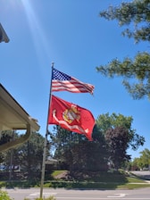 Two flags are flying on flagpoles against a clear blue sky. The American flag is at the top, and below it is a red United States Marine Corps flag. Pine trees are visible in the background, along with a street and a part of a residential house roofline.