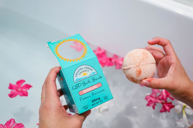 A hand holds a blue box labeled 'CBD Bath Bomb' with the words 'Sensual Passion Romance.' Another hand holds a round, light orange bath bomb. Pink flowers float on the water in the background, partially visible in the tub.