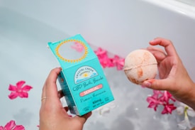 A hand holds a blue box labeled 'CBD Bath Bomb' with the words 'Sensual Passion Romance.' Another hand holds a round, light orange bath bomb. Pink flowers float on the water in the background, partially visible in the tub.