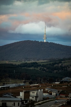 A telecommunications tower stands prominently on a tree-covered hill under a sky filled with dramatic clouds. Below, modern residential houses are scattered across a lush, green landscape.