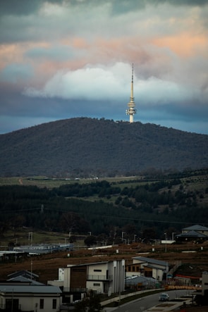 A telecommunications tower stands prominently on a tree-covered hill under a sky filled with dramatic clouds. Below, modern residential houses are scattered across a lush, green landscape.