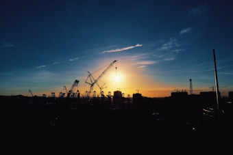 A silhouetted construction site featuring several large cranes stands against the backdrop of a stunning sunset. The sky is painted with a gradient of deep blue transitioning into warm orange and yellow near the horizon, creating a striking contrast with the dark outlines of the cranes and buildings.