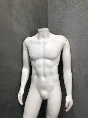 A headless white mannequin torso stands upright against a textured gray wall, displaying a muscular physique.