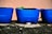 Three blue plastic containers, likely repurposed as plant pots, sit on a rough, textured surface. Sparse green plants or seedlings are emerging from the soil in the middle pot. The background consists of a muted pink wall.