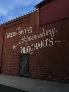 A brick wall with vintage-style painted signage for a store named TINKER SMITHS, advertising services like haberdashery and merchants. The colors are muted, with a focus on the contrast between the brick red and white lettering. A wooden door is also present on the wall.
