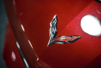 A close-up view of a car badge featuring a silver and black checkered flag design and a red emblem with a Chevrolet logo set against a metallic red car surface. The surface reflects light, creating bright highlights.