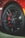 A close-up view of a black alloy wheel with a red brake caliper. The wheel has a unique design with multiple spokes and a branded logo at its center. The tire is visible, illustrating its tread pattern.