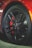 A close-up view of a black alloy wheel with a red brake caliper. The wheel has a unique design with multiple spokes and a branded logo at its center. The tire is visible, illustrating its tread pattern.