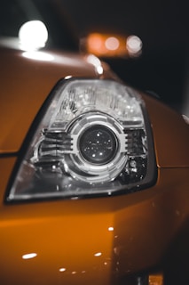 A close-up shot of a car's headlight, featuring intricate details of the glass and metallic components. The headlight is part of an orange vehicle with reflections of light visible on the surface.