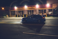 A blue car is driving on a dimly lit street at night. The scene is in front of a building with illuminated signage, indicating a business called North Atlantic Supplies Inc.