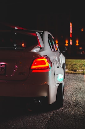 A white sports car parked on an illuminated street at night, featuring a rear spoiler and glowing red tail lights. The background includes blurred lights from nearby buildings, adding a city atmosphere.