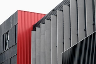 A modern architectural facade composed of geometric panels, primarily featuring a combination of red, gray, and black colors. The design features vertical lines and a mix of textures.